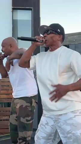 The video shows two men performing on a stage outdoors. The man in the foreground is wearing a white t-shirt, sunglasses, and a black baseball cap. He is holding a microphone and gesturing with his left arm. He is also wearing camouflage pants. The man in the background is wearing a white t-shirt and camouflage pants. He has his arm around the man in the foreground. The background shows a building with large windows and some wooden structures. The lighting suggests it is daytime.