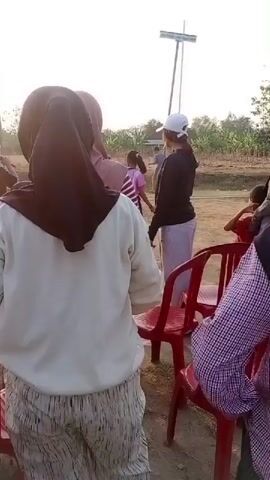A group of people, mostly women wearing hijabs, are gathered outdoors. In the foreground, a woman in a white sweatshirt and patterned pants is visible from behind, facing away from the camera. To her left, another woman in a pink hijab is also seen from behind. To the right, several red plastic chairs are arranged, with people sitting and standing around them. In the background, more people, including some children, are present, and there are trees and an open field. A tall, thin structure, possibly a utility pole or flag pole, is visible in the distance against a bright sky. The lighting suggests it is either early morning or late afternoon. The overall atmosphere appears to be a casual gathering or event.