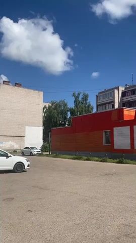 The video shows a residential area on a sunny day. It pans across a street with parked cars, apartment buildings, trees, and a blue sky with white clouds. There are no people visible in the video. The buildings are made of brick and concrete, and some have air conditioning units. There is a mural on the side of one building. The overall impression is of a quiet, everyday urban environment.