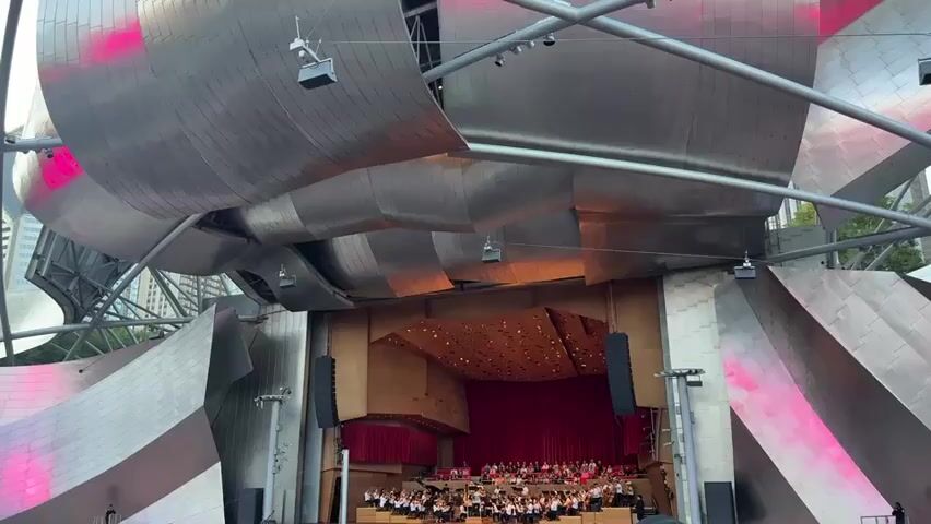 An exterior shot of the Jay Pritzker Pavilion, an outdoor amphitheater in Chicago, shows its distinctive wavy metal roof structure. A symphony orchestra is seated on the stage, with a red curtain behind them. Pink lights illuminate parts of the metal roof. The sky is clear blue.
