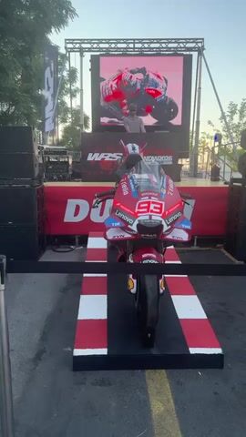 A promotional event features a red and white Ducati motorcycle with the number 93 on display. Behind the motorcycle is a stage with a large LED screen showing racing footage, and a DJ is visible on the stage. The motorcycle is on a display ramp with red and white checkered accents. The event appears to be outdoors, with trees and a clear sky in the background.