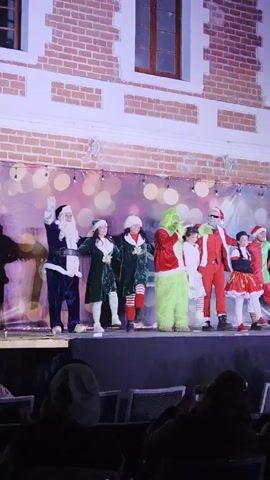 The video shows a group of people dressed in Christmas-themed costumes waving to an audience. The individuals on stage appear to be dressed as Santa Claus, elves, and the Grinch. The backdrop features a decorative wall with Christmas lights, which suggests a holiday event or performance. The audience is seated and watching the show. The setting seems to be an outdoor stage.