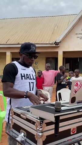 A man wearing a black baseball cap with a white “A”, sunglasses, a white basketball jersey with the name “HALL” and the number “17” on it, and a black t-shirt underneath is operating a DJ mixer.  The mixer is in a silver metal case with a microphone next to it on the case. A laptop is open on the case as well, displaying the words "DJ Blaze" and a website address. A group of children stand behind the DJ looking at him.  They are in a courtyard outside a single-story building with a tan roof and brown trim. A sign on the wall of the building is partly visible, and a green plastic chair is visible.