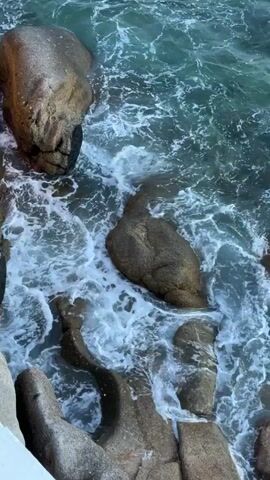 A high-angle shot captures ocean waves crashing against large, weathered rocks. The water is a deep teal with white foam from the impact of the waves. Some of the rocks are partially submerged, while others are exposed and rounded. The scene conveys the raw power and movement of the ocean.