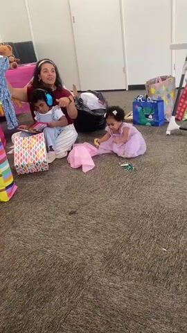 A woman is sitting on the floor with two young children. She is holding a blue garment above the head of one child who is wearing blue and white clothing and noise-canceling headphones. The other child, dressed in a lavender dress, is crawling on the carpet near a pile of pink tissue paper and a green object. There are several gift bags around them, including a pink one with a teddy bear and a blue one with a dinosaur. The environment appears to be indoors with light-colored walls and doors in the background.