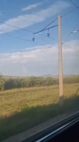 The video shows a view from a moving vehicle, likely a train, looking out a window. The landscape consists of a grassy field with some scattered trees and bushes. In the distance, there appear to be rolling hills or mountains. The sky is blue with some white clouds, and power lines are visible overhead. The lower portion of the frame shows the window frame of the vehicle and a hint of the tracks or roadbed. The lighting suggests it is daytime, possibly late afternoon or early morning given the angle of the sun and the shadows. There are no people or animals visible in the frame. The scene is predominantly rural.