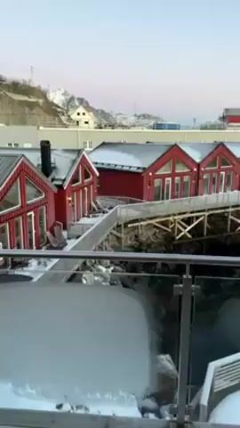 The camera pans left to show a row of red wooden cabins with white trim on a harbor in a snowy, mountainous landscape. The roofs of the cabins are covered in snow, and they are connected by wooden walkways. There is a small dock covered in snow in the water in front of the cabins. The water is calm, and there are snow-covered mountains in the distance. The sky is clear and blue, with a hint of pink on the horizon. The camera is positioned behind a glass railing.