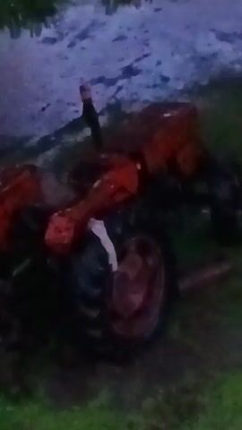 The video shows a red tractor on a grassy area, with what appears to be a person standing on or near the tractor.  The lighting is poor, making details difficult to discern. There's a muddy or whitish area in the background. It's difficult to tell the exact condition of the tractor or what the person is doing.