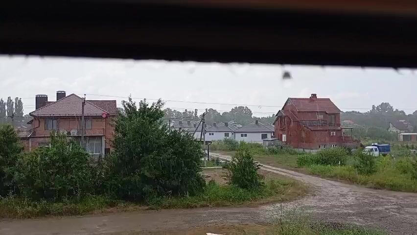 The video shows a view through a window on a rainy day. Water droplets are visible on the glass, obscuring the view of an outdoor scene. The outdoor scene depicts several houses, some with red tile roofs, and green trees. A dirt road or path is visible in the foreground, leading towards the houses. The overall atmosphere is overcast and gloomy due to the rain.