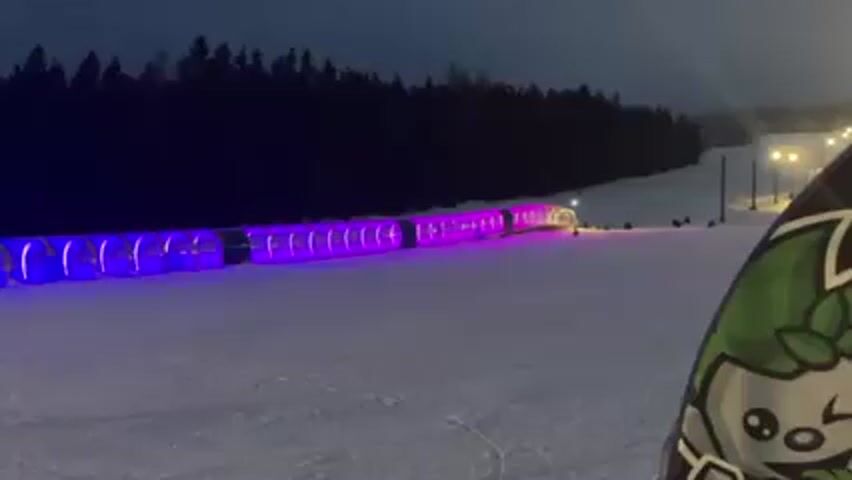 The video shows a winter sports area at night. A snow-covered slope is illuminated by streetlights and a long tunnel made of connected, translucent sections. The sections of the tunnel cycle through various colors, including blue, green, yellow, red, and purple. The edge of a snowboard with a cartoon logo is visible in the foreground. Several figures can be seen scattered on the slope near the tunnel exit. A dark treeline is silhouetted against the night sky.