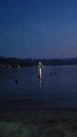 The video shows a body of water at dusk. In the distance, there is a boat with lights on. There are several buoys in the water closer to the foreground. The sky is a deep blue with some faint clouds. Hills are visible on the horizon. The foreground shows a sandy or rocky beach area. The overall scene appears calm and serene.