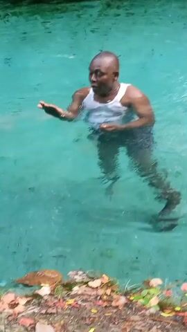 A man is standing in a clear blue body of water. He is wearing a white tank top and dark shorts. He is smiling and gesturing with his hands. The water is shallow enough that he is standing with his feet on the bottom. The edge of the water is lined with plants and leaves.