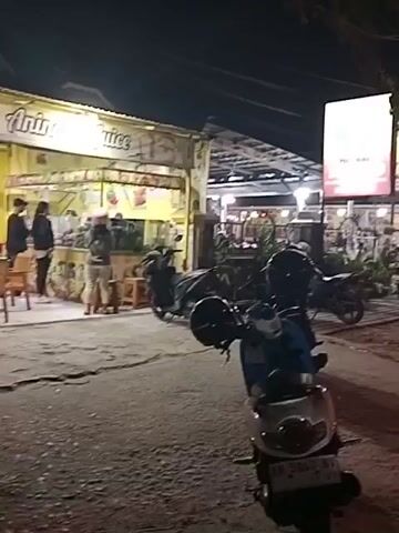 The video shows a nighttime street scene. In the foreground, a parked scooter is visible. Behind it, several more scooters are parked. To the left, a food stall with a "Juice" sign is illuminated. Further back, a row of buildings with open fronts and lights suggests a commercial area. People can be seen standing near the food stall. The street itself is paved and appears wet, reflecting the lights. Traffic, including cars and motorcycles, is moving along the road in the background. The overall atmosphere is that of a busy street at night, likely in a tropical or subtropical region given the presence of scooters and open-air establishments.
