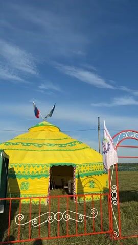 A yellow yurt with green trim and symbols stands in a grassy field under a blue sky with white clouds. Two flags, one red, white, and blue, and another green, fly from the top of the yurt. In the foreground, a red metal archway with white decorative swirls stands over a red metal fence. To the right of the archway is a sign with green and white lettering and a logo featuring a gear. Another green structure is partially visible to the left of the yurt. The scene appears to be outdoors, possibly at an event or festival.