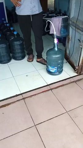 A person stands in front of a large plastic water jug that is being filled from another jug. The person is wearing black pants and a grey t-shirt and is standing with their left hand on their hip. The filling jug is a clear plastic jug with a blue lid. The jug being filled is a large, dark blue plastic jug with a white label that says "AQUA". A white hose is connected to the top of the filling jug and extends down into the jug being filled. The scene appears to be a shop or warehouse, with many other large plastic water jugs stacked in the background. The floor is tiled with white tiles.