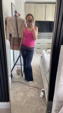 A young woman with brown hair tied back in a ponytail stands in front of a full-length mirror, taking a selfie. She is wearing a bright pink tank top and dark blue flared pants with a striped waistband. Her nails are painted a bright pink. She poses with her arms bent and her hands near her head. In the background, a clothing rack with a beige and brown plaid garment is visible, as is a white fan and some charging cables on the floor. A bed with white bedding is to the right of the mirror. The room appears to be a bedroom, with wooden closets and a TV visible in the background.