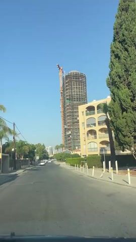 The video shows a street view with a tall, cylindrical building under construction in the background. A crane is visible attached to the side of the building. To the right of the construction, there is a beige apartment building with balconies. The street is paved and there are cars parked on the left side. There are trees and bushes lining the street. The sky is clear and blue. The perspective suggests the video was recorded from inside a vehicle.