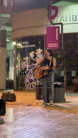 A man with dark, curly hair and a mustache stands in front of a glass-fronted building, playing an acoustic guitar and singing. He is wearing a black tank top, grey pants, and white sneakers. A microphone is positioned in front of him. To his left, on the ground, is a black speaker. To his right, behind him, is a large cylindrical trash receptacle. Above and behind him, a sign with the word "ISTANBUL" is visible, along with a purple banner that reads "ibb.kultur sahnem istanbul". Inside the glass building, a large illustration of a person riding a bicycle is visible. The ground is paved with cobblestones. Another person walks past in the background to the right.