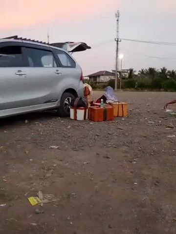 A silver minivan with its tailgate open is parked on a gravelly surface. Several rectangular packages, some wrapped in brown paper and tied with twine, are arranged on the ground next to the open tailgate. One package appears to be a basket with a lid. A person wearing a headscarf is seated next to the packages. In the background, there is a structure that appears to be a house, a utility pole with wires, and some vegetation. To the right, another person is seated on the ground, facing away from the camera. The sky is overcast and appears to be dusk.