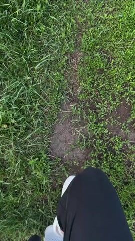 The video begins with a view of a grassy path from the perspective of someone walking, with a white sneaker visible in the lower part of the frame. The camera then lifts to reveal a vast green field under a blue sky with white clouds. The camera pans across the field, showing sparse trees and a distant treeline. The overall scene is a rural, open landscape.