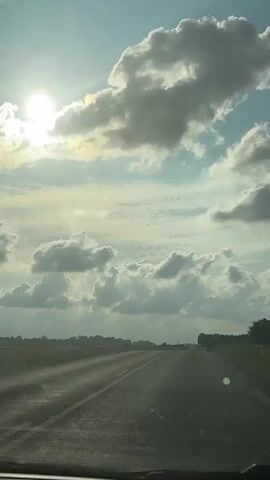 The video shows a view from a car moving on a road. The sun is shining brightly in the sky, partially obscured by clouds. The road stretches ahead, lined by fields on either side. The scene appears to be in a rural or semi-rural area. There are no people visible in the frame. The time displayed is 00:00 to 00:04.