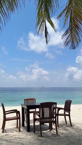 The video depicts a tranquil beach scene. A black table with four wooden chairs sits on white sand, positioned to face the ocean. The ocean is turquoise near the shore, gradually darkening to blue further out.  The sky above is bright blue, dotted with fluffy white clouds. Palm tree fronds overhang the scene, providing partial shade. In the distance, two small islands are visible on the horizon. No people are visible in the shot. The overall atmosphere is peaceful and serene.