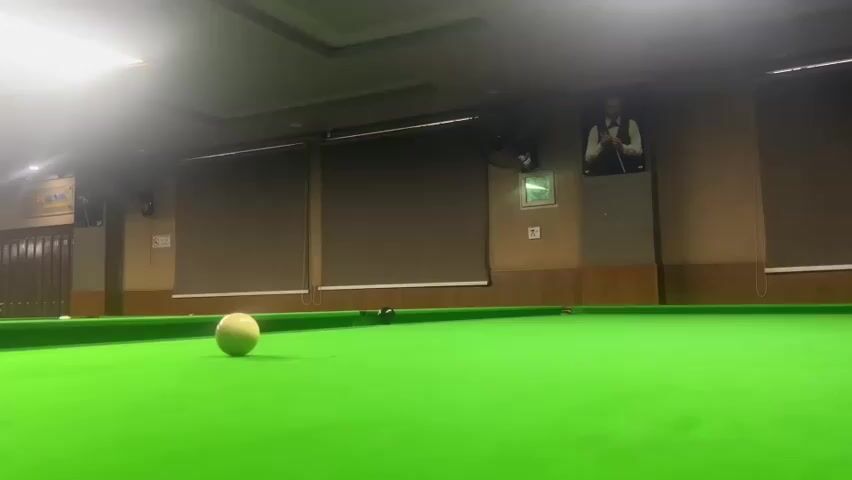 A low-angle shot captures a close-up view of a snooker table, with a white cue ball positioned on the green felt surface. In the background, a man wearing a suit and tie is visible, holding a snooker cue in his hands, suggesting he is about to play. The room is dimly lit, with overhead lights casting a warm glow on the table. The walls are paneled with wood, and several windows are covered with dark blinds. The overall atmosphere appears to be that of a snooker club or a private room set up for the game.
