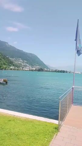 A fixed, wide shot captures a serene waterfront view on a clear, sunny day. The foreground is dominated by a wooden deck area with a metallic railing and a wooden bench. A dark flag with a turquoise logo flutters from a flagpole positioned on the right side of the deck. Beyond the deck, a vast expanse of blue water stretches out, leading to a distant shoreline lined with buildings and rolling, green-covered mountains under a bright blue sky. A small boat is visible on the water in the middle ground. The overall impression is one of a tranquil, scenic outdoor location, possibly a resort or a park by a lake.