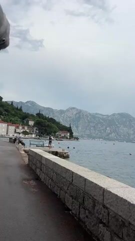 This is a video clip of a man standing on a stone pier by the sea, with a view of mountains and buildings in the background. The setting appears to be Perast, Montenegro. The man is wearing a t-shirt and shorts and a blue baseball cap. He is facing the water and appears to be getting ready to enter it or has just exited. The weather seems overcast, with a cloudy sky. There are boats in the water and buildings lining the shore. In the foreground, there is a paved path and a stone wall. Two park benches are also visible on the pier. The overall mood is relaxed and leisurely, suggesting a warm day for swimming. No visible text is present.