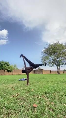 A shirtless Black man is performing a handstand on a grassy field on a sunny day. He is wearing black pants and shoes. There is a blue jacket lying on the grass near him. Behind him, there is a brick wall and several trees. The sky is blue with scattered white clouds.
