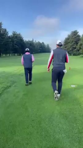 Two golfers are walking down the fairway of a golf course on a sunny day. The golfer on the left is wearing a pink vest, navy pants, and a white hat. The golfer on the right is wearing a navy vest with red and white accents, navy pants, and a white hat. They are walking away from the camera. The fairway is green and well-maintained, with trees lining the sides. The sky is blue with some clouds. There is a red tee marker on the right side of the fairway.