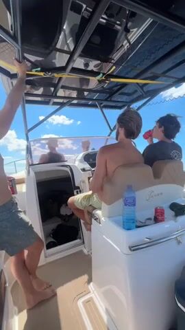 The video shows four young men on a boat on a sunny day. The camera is positioned inside the boat, looking out towards the front and side. One man is standing on the left, holding onto a metal bar. Another man is in the driver's seat, wearing a checkered shirt. Two other men are visible in the background, one in the passenger seat and another further back. The boat appears to be moving, with water splashing up on the side. The sky is blue with white clouds, and the ocean is visible in the distance.