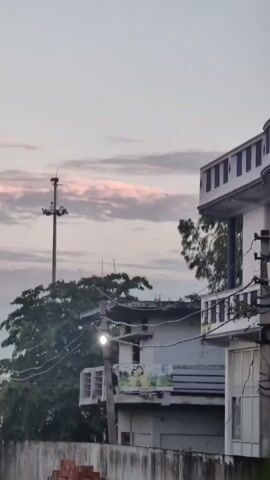 The video pans from a low angle upwards to a cloudy sky. In the foreground, a concrete wall runs horizontally across the bottom of the frame. To the left, a utility pole with wires is visible. Further back, a building with a white facade and a balcony can be seen, with trees and power lines in front of it. A tall, narrow tower with lights on top is situated in the distance. The sky displays soft clouds with hints of pink and grey. The scene appears to be an urban or suburban environment during twilight or dawn.