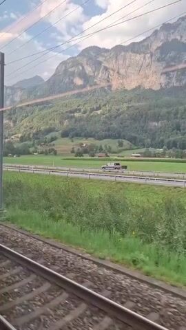 This video is a sideways-on, medium-shot, point-of-view recording from a moving train. The train tracks are visible in the foreground, with grass and vegetation on the right. A highway runs horizontally through the middle ground, with a white car visible on the left side of the highway and a light-colored pickup truck towing a trailer visible in the center. Beyond the highway, a green field slopes upwards toward a dense forest that covers the lower part of a large, rugged mountain range. The mountain range is light brown and grey, with patches of green vegetation. The sky above is partly cloudy with blue sky visible. The overall scene is pastoral and mountainous.