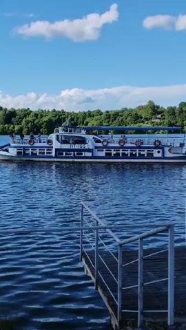 This media file features a wide shot of a river on a clear, sunny day, with a passenger boat in the mid-ground. The boat is white with a blue stripe along its side and has several life preservers visible. People can be seen on the boat's upper deck. The water is blue and slightly rippled, suggesting a gentle breeze. In the foreground, a wooden pier with a metal railing extends into the water from the right side of the frame. The background consists of a lush green treeline with some houses nestled among the trees, indicating a riverside settlement. The sky is a vibrant blue with scattered white, puffy clouds. The scene conveys a sense of leisure and a pleasant summer day. The text "ПТ-165" is visible on the side of the boat. The location context provided is Plyos, Russia.