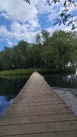 A long, straight wooden pier extends from the foreground over dark, reflective water towards a distant landmass covered in lush green trees. The water mirrors the trees and parts of the blue, partly cloudy sky. The sky above features scattered white clouds against a bright blue background. The pier, made of weathered wooden planks, appears to be the sole path across the water. The trees on the far side form a dense green line, suggesting a wooded area or an island. The scene is tranquil and appears to be set during daytime under clear weather conditions in Kipen, Russia. No people are visible in the video.