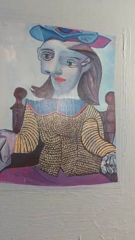 A close-up video shows a printed image of a cubist-style painting of a woman wearing a blue hat and a patterned shirt. The painting is taped to a textured, light-colored wall. The camera slowly zooms in on the painting, revealing details of the artwork. The woman in the painting has a fragmented face, with two distinct profile views of her face overlaid on each other. She is seated in a chair, with her hands resting in her lap. Her clothing is depicted in vibrant colors and intricate patterns. The background of the painting is a light blue, possibly representing the sky or a wall. The video ends with a final close-up view of the woman's face.
