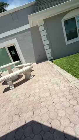 The video pans across a paved patio area with a stone bench and table. A bird is seen walking on the grass next to the patio. The house is painted gray with white trim. The sky is visible in the background.