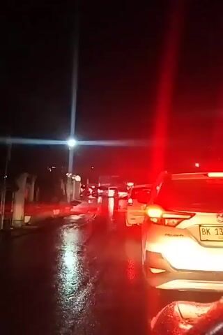 The video shows a nighttime traffic jam on a wet road. Cars are stopped, with their brake lights illuminated, creating red reflections on the wet asphalt. The overall lighting is dim, with some streetlights and vehicle headlights visible. The perspective is from inside a vehicle, looking forward.
