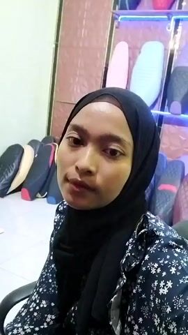 A young woman wearing a black hijab and a dark floral print shirt looks into the camera. She is seated in a black chair, and behind her, various motorcycle seats in different colors and styles are displayed on shelves. The lighting appears to be artificial, with some blue neon lights visible on the shelves behind the woman.
