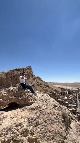 A man sits on a large rock overlooking a town nestled in a valley under a bright blue sky. He appears to be taking photos with his phone. Later, a second man in a dark jacket and hat stands at the same spot, looking out over the town and holding a water bottle. The town is composed of buildings with light-colored roofs and is surrounded by a semi-arid landscape. A road runs through the town. The rock the men are on is light brown and rugged. The sky is mostly clear with a few small clouds.