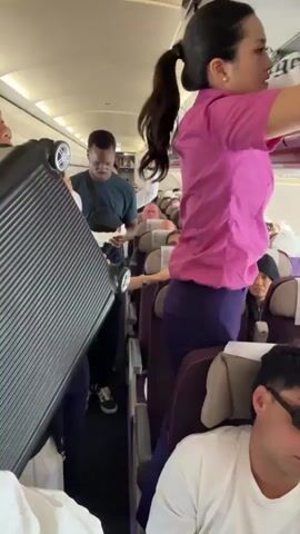 The interior of an airplane cabin is captured in this video. Several people are present, including two female flight attendants wearing pink shirts and dark purple pants, who appear to be assisting passengers with their luggage. One flight attendant is standing on a seat, reaching into an overhead compartment to place a bag. Another flight attendant holds a large, black suitcase for a passenger.  The passengers seated in the foreground include a man wearing sunglasses and a white t-shirt with writing on it, and a young girl looking directly at the camera. The background shows other passengers seated, and some standing in the aisle. The lighting inside the cabin is bright, and the interior is clean and well-maintained.