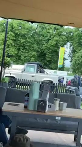 Here is a description of the video:

The video shows a view from under a large outdoor umbrella, probably on a deck or patio. There are tables and outdoor seating, with gray cushions. There is a mug on the table in the foreground.  Beyond the seating area, there's a low black fence, and beyond that, a large grassy area with trees and what appears to be an event taking place. There are structures and people walking around in the distance. There is also a white pick-up truck visible behind the fence in the first few seconds of the video. The sky is overcast.