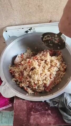 A close-up overhead shot shows a large metal pot filled with cooked rice and chopped carrots. A silver spoon is seen in the pot, stirring the contents. The pot sits on what appears to be a white enamel stove with some dark marks on its surface. The background shows a rough, textured surface, possibly a floor or countertop, and a hint of a pink fabric to the left and a gray cloth to the right. The lighting is somewhat dim, creating shadows around the pot.