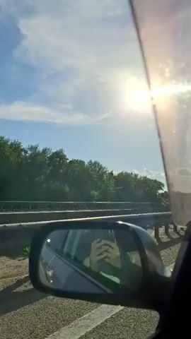 The video shows a view from inside a car, looking out the side window. The sun is bright and shining in the sky, with some clouds visible. Trees and a guardrail are seen along the side of the road. In the side mirror, a person can be seen holding a phone, presumably taking a selfie or video. A truck is briefly visible in the distance. The scene appears to be a car driving on a highway on a sunny day.