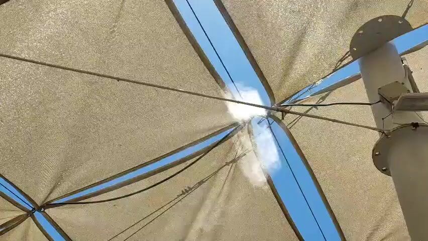 A low-angle shot of a beige fabric shade structure with blue sky visible through gaps. A mist is sprayed from a nozzle located at the center of the structure. The mist drifts away from the nozzle.