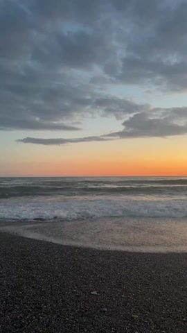 The video shows a beach at sunset. The sky is filled with gray clouds, but the horizon is a bright orange. The ocean waves are crashing on the shore, which is covered with small, dark pebbles. There are no people visible in the video.