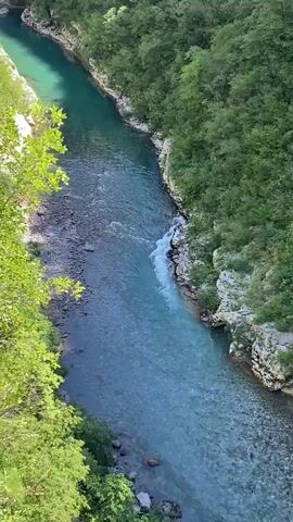 The video captures a serene aerial view of a vibrant turquoise river winding through a lush green canyon. The river, with its exceptionally clear water, reveals rocky beds beneath its surface. On the left bank, the shore is a mix of darker rocks and light green foliage, while the right bank is characterized by rocky outcrops and dense, dark green trees. In the mid-ground, the river creates a small section of foamy rapids as it flows over submerged rocks, contrasting with the calmer, deeper sections. The scene suggests a sunny day with excellent visibility, highlighting the natural beauty of the river and its surrounding forested gorge. No people or text are visible in the video.