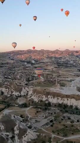 A panoramic aerial view captures numerous hot air balloons in various colors and sizes ascending into a clear, pale blue sky at sunrise. The balloons are scattered across the frame, some in the foreground and others dotting the distant horizon. Below, a vast landscape unfolds, characterized by unique geological formations resembling fairy chimneys, rocky outcrops, and valleys. Patches of green vegetation and what appear to be agricultural fields are visible between the rock formations. Buildings, likely part of a settlement, are nestled within the landscape in the mid-ground. The soft, warm light of dawn casts gentle shadows across the terrain, highlighting its textured surface. The overall scene conveys a sense of peacefulness and wonder.