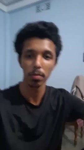 A close-up, slightly blurry video shows a young man from the chest up. He has dark, curly hair and a mustache. He is wearing a black t-shirt. The background is a light blue wall. There is a blurred object, possibly a towel, hanging on the wall to the right. The man's head is tilted slightly and he is looking towards the camera. His expression is neutral.